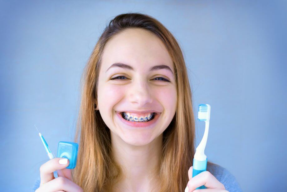 Young girl with long straight light brown hair smiling with braces while holding a tooth brush and floss.