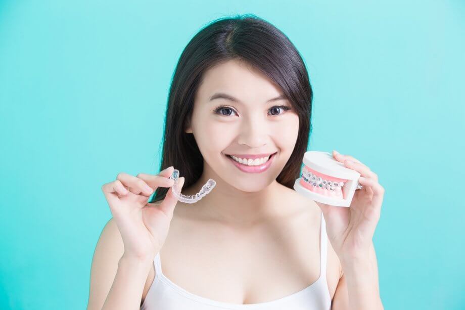 Photograph of a woman with brown hair holding Invisalign braces in one hand & traditional braces on false teeth in the other.